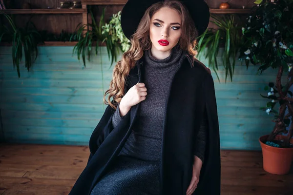 Молодая красивая модная леди сидит у большого окна в кафе и держит чашку кофе. Привлекательная женщина в стильной черной шляпе . — стоковое фото
