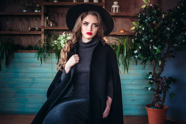 Młoda piękna Pani modne siedzi w pobliżu duże okno w kawiarni i trzymając kubek kawy. Atrakcyjna kobieta sobie stylowy kapelusz czarny. — Zdjęcie stockowe