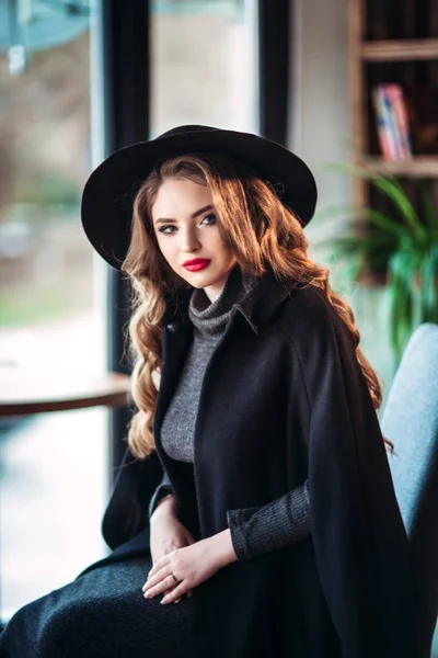 Joven hermosa dama de moda sentada cerca de la ventana grande en la cafetería y la celebración de la taza de café. Mujer atractiva con sombrero negro con estilo . —  Fotos de Stock