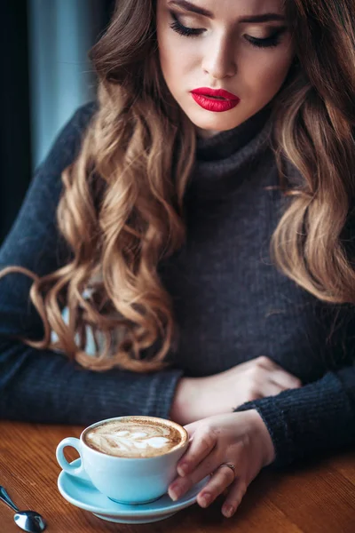 Beautiful young woman enjoying coffee cappuccino with foam near window in a cafe — Stock Photo, Image