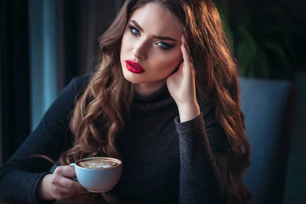 Hermosa joven mujer disfrutando de café capuchino con espuma cerca de la ventana en un café — Foto de Stock