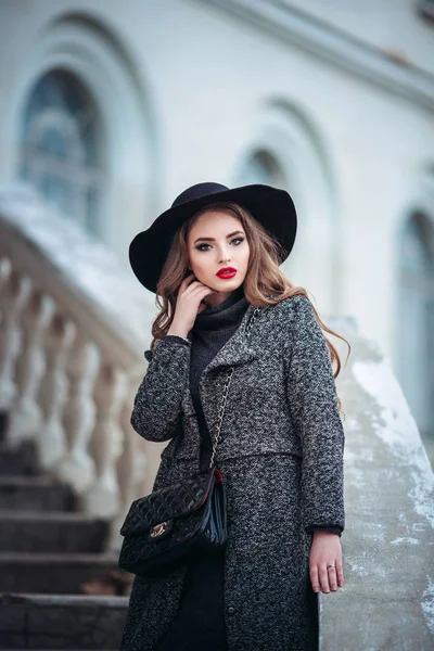 Joven chica hermosa con maquillaje perfecto, labios rojos, con un sombrero negro y abrigo gris, vestido negro, posando en evning city . —  Fotos de Stock