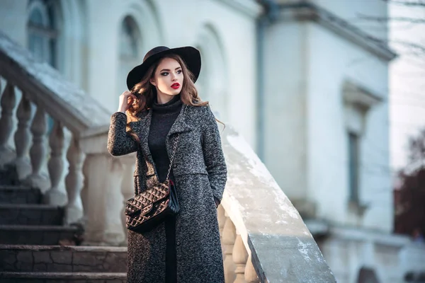 Junges schönes Mädchen mit perfektem Make-up, roten Lippen, trägt einen schwarzen Hut und grauen Mantel, schwarzes Kleid, posiert in der nächtlichen Stadt. — Stockfoto