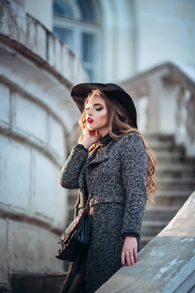 Joven chica hermosa con maquillaje perfecto, labios rojos, con un sombrero negro y abrigo gris, vestido negro, posando en evning city . —  Fotos de Stock