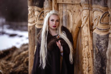 Kürk yakalı bir geleneksel giysiler içinde güzel öfkeli İskandinav savaşçı zencefil kadınla kılıç onun el ve ahşap Viking köy görünümünde arka planda açık havada portresi.