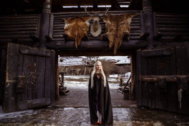 Kürk yakalı bir geleneksel giysiler içinde güzel öfkeli İskandinav savaşçı zencefil kadınla kılıç onun el ve ahşap Viking köy görünümünde arka planda açık havada portresi.