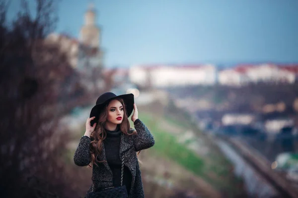 Joven chica hermosa con maquillaje perfecto, labios rojos, con un sombrero negro y abrigo gris, vestido negro, posando en evning city . — Foto de Stock