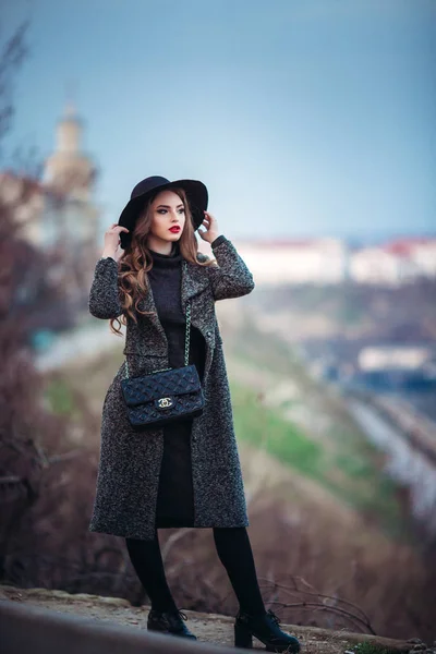 Joven chica hermosa con maquillaje perfecto, labios rojos, con un sombrero negro y abrigo gris, vestido negro, posando en evning city . — Foto de Stock