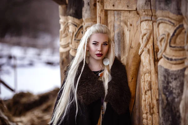 Outdoors portrait of beautiful furious scandinavian warrior ginger woman in a traditional clothes with fur collar, with sword in her hand and wooden Viking Village view on the background. — Stock Photo, Image