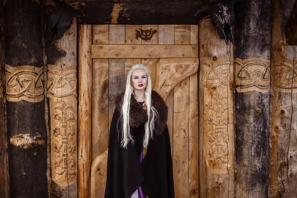 Outdoors portrait of beautiful furious scandinavian warrior ginger woman in a traditional clothes with fur collar, with sword in her hand and wooden Viking Village view on the background. — Stock Photo, Image