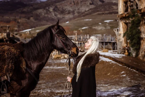 Outdoors portrait of beautiful furious scandinavian warrior ginger woman in a traditional clothes with fur collar, with sword in her hand and wooden Viking Village view on the background. — Stock Photo, Image