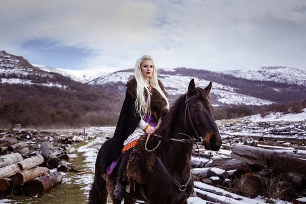 Outdoors portrait of beautiful furious scandinavian warrior ginger woman in a traditional clothes with fur collar, with sword in her hand and wooden Viking Village view on the background. — Stock Photo, Image