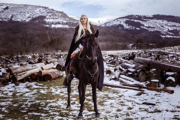 Outdoors portrait of beautiful furious scandinavian warrior ginger woman in a traditional clothes with fur collar, with sword in her hand and wooden Viking Village view on the background. — Stock Photo, Image