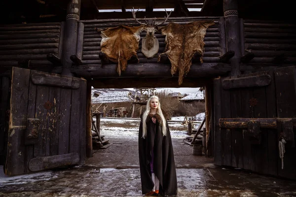 Ao ar livre retrato da bela guerreira escandinava furiosa mulher gengibre em uma roupa tradicional com gola de pele, com espada na mão e vista Viking Village de madeira no fundo . — Fotografia de Stock