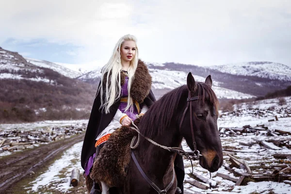 Outdoors portrait of beautiful furious scandinavian warrior ginger woman in a traditional clothes with fur collar, with sword in her hand and wooden Viking Village view on the background. — Stock Photo, Image