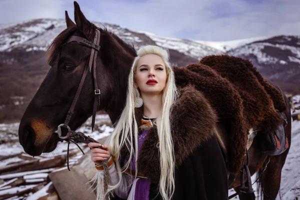 Buitenshuis portret van mooie woedend Scandinavische warrior gember vrouw in een traditionele kleding met bont kraag, met zwaard in haar hand en houten Viking Village bekijken op de achtergrond. — Stockfoto