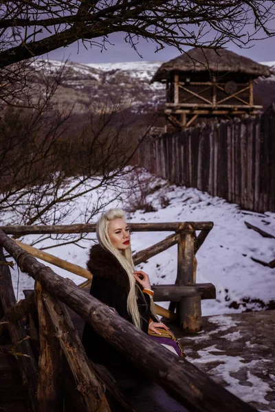 彼女の手と背景に木製のバイキング ビレッジ ビューで剣と毛皮の襟付きの伝統的な服の美しいワイルド北欧戦士の生姜女性の屋外ポートレート. — ストック写真