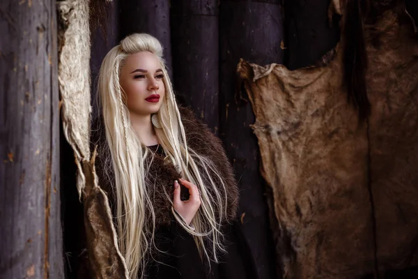 На улице портрет красивой яростной скандинавской воительницы рыжей женщины в традиционной одежде с меховым воротником, с мечом в руке и деревянным видом на деревню викингов на заднем плане . — стоковое фото