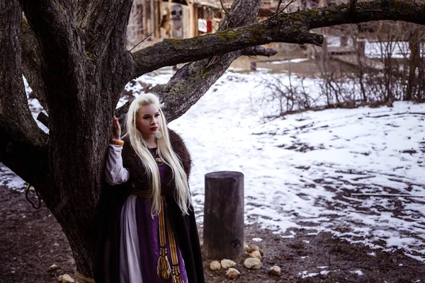 Buitenshuis portret van mooie woedend Scandinavische warrior gember vrouw in een traditionele kleding met bont kraag, met zwaard in haar hand en houten Viking Village bekijken op de achtergrond. — Stockfoto