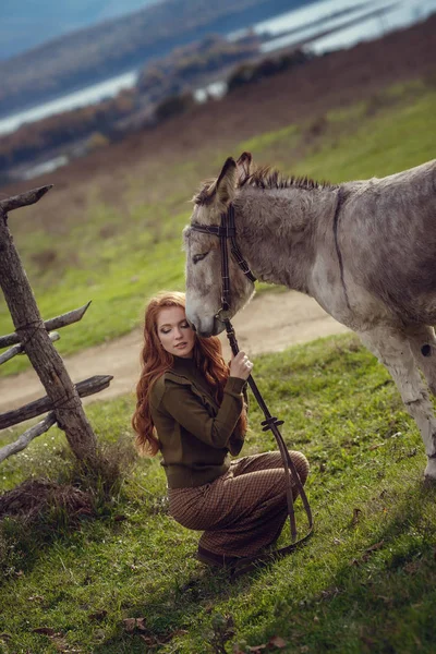 A girl with curly red hair in fashionable clothes in the style of Provence hugs a cute donkey — Stock Photo, Image