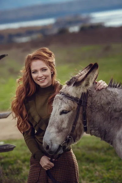 A girl with curly red hair in fashionable clothes in the style of Provence hugs a cute donkey — Stock Photo, Image