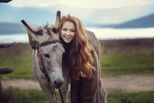 Een meisje met krullend rood haar in modieuze kleding in de stijl van de Provence hugs een schattig ezel — Stockfoto