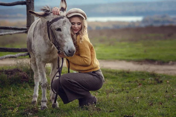 Ein Mädchen mit blonden Haaren in modischer Kleidung im Stil der Provence umarmt einen niedlichen Esel — Stockfoto