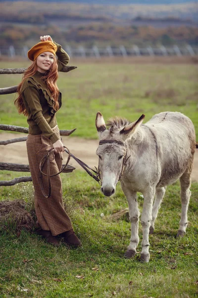 一个卷曲的红头发女孩穿着时髦的衣服, 以普罗旺斯的风格拥抱了一头可爱的驴 — 图库照片