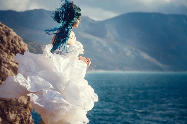 Молода жінка стоїть на краю скелі і дивиться на широкий вид на море. Вітер Довга блакитна зачіска . — стокове фото