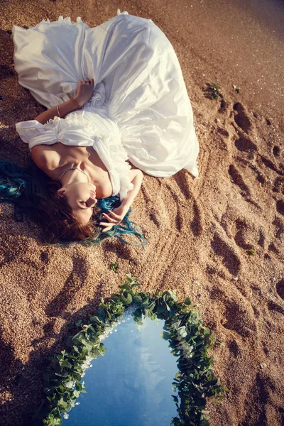 Mooi meisje met blauwe haren en in een witte vintage jurk kijkt naar de zee. In haar handen een mooie antieke spiegel, waarin de zee weerspiegelt. Boekomslag — Stockfoto