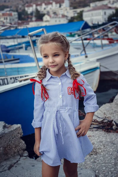 Krásná holčička v mořském stylu na pozadí člunů a jachtů. Malý námořník — Stock fotografie