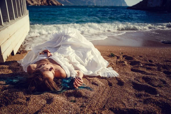 Hermosa chica con el pelo azul y en un vestido vintage blanco mira al mar. En sus manos un hermoso espejo antiguo, en el que el mar se refleja. Portada del libro Imagen De Stock