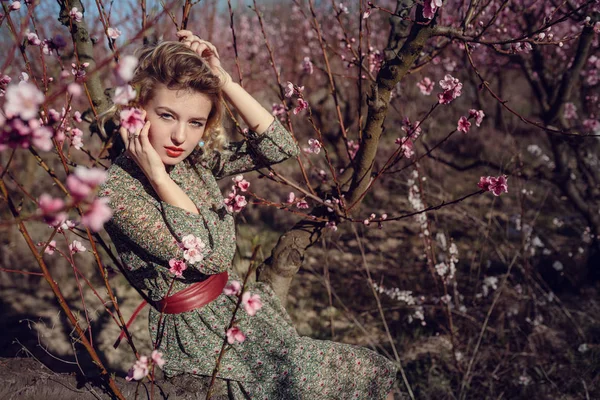 Модная открытая фотография великолепной молодой женщины в элегантном платье позирует в саду с цветущими персиковыми деревьями — стоковое фото