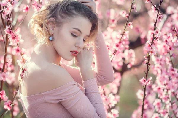 Foto de moda al aire libre de la hermosa mujer joven en vestido elegante posando en el jardín con árboles de melocotón en flor —  Fotos de Stock