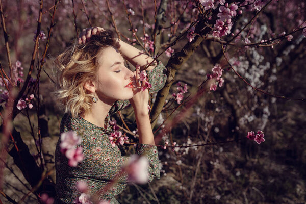Fashion outdoor photo of gorgeous young woman in elegant dress posing in garden with blossom peach trees. Blonde in flowering gardens