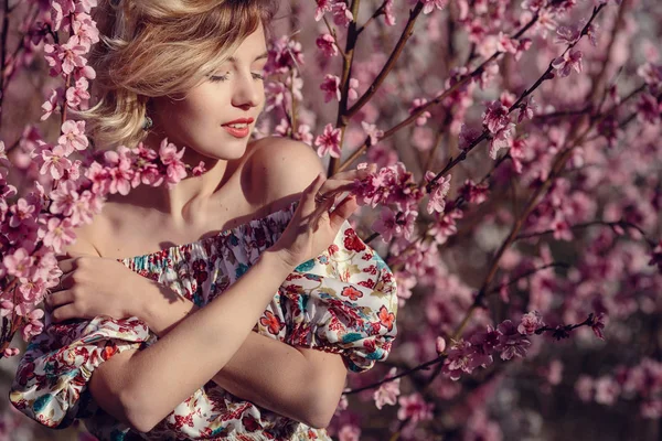 Mode buiten foto van prachtige jonge vrouw in elegante jurk poseren in de tuin met perzik bloesem bomen — Stockfoto