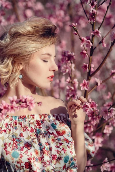 Mode buiten foto van prachtige jonge vrouw in elegante jurk poseren in de tuin met perzik bloesem bomen — Stockfoto