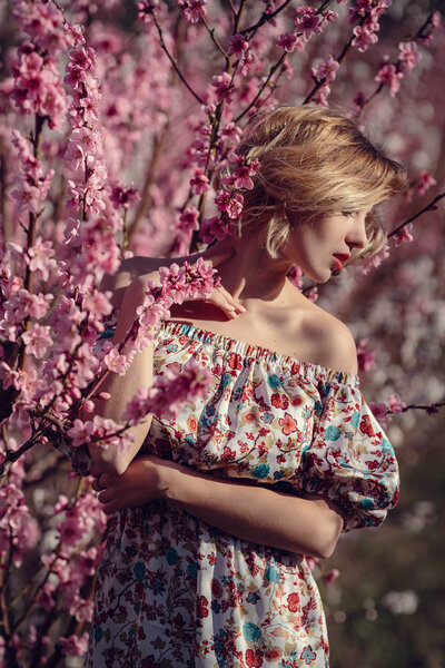 Fashion outdoor photo of gorgeous young woman in elegant dress posing in garden with blossom peach trees. Blonde in flowering gardens