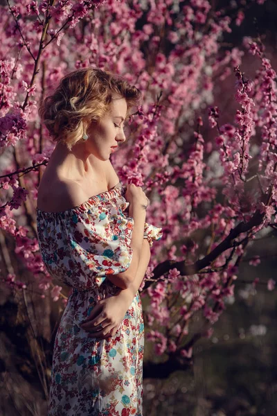 Mode buiten foto van prachtige jonge vrouw in elegante jurk poseren in de tuin met perzik bloesem bomen — Stockfoto