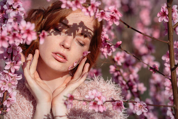 Beautiful young girl with red hair in a gentle peach garden, which blossomed. The concept and idea of skin and hair care.