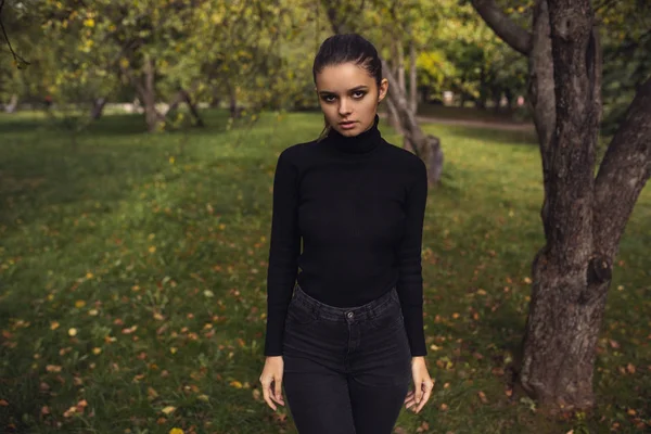 A beautiful young brunette walks through the autumn park in a black sweater and coat, — Stock Photo, Image