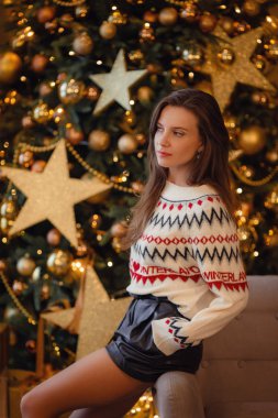 Beyaz kazak giyen genç ve güzel bir kadın yeni yıl ağacının yanında oturuyor.