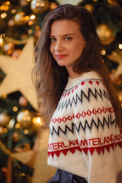 Uma bela jovem mulher em uma camisola branca está sentada perto da árvore de Ano Novo — Fotografia de Stock