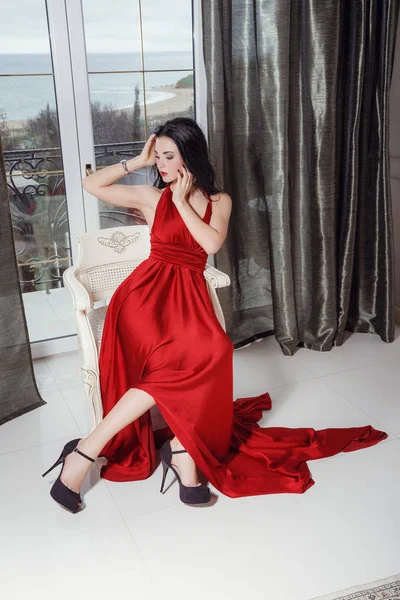 Sexy půvabná žena s černými vlasy v elegantních červených šatech sedícího na křesle — Stock fotografie