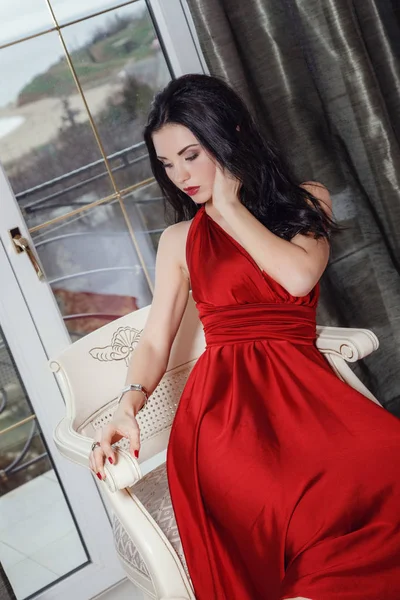 Sexy půvabná žena s černými vlasy v elegantních červených šatech sedícího na křesle — Stock fotografie