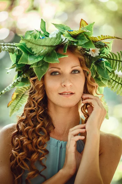 a beautiful young woman with red hair and a wreath of flowers and fern in the forest celebrates Solstice Day, mid-summer.