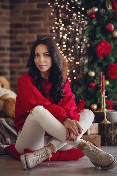 Beautiful elegant woman with wavy hair and makeup in a red sweater and white jeans posing in dark evening xmas decorated interior with christmas tree and lights. Cozy portrait