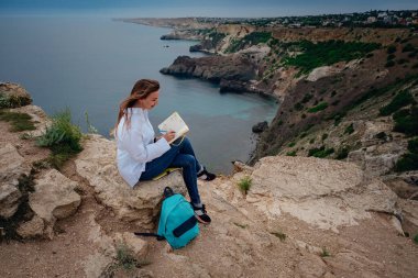 Çekici bir kadın, deniz manzaralı bir dağda oturmuş kitap okuyor. Açık havada kendini eğitmek. günlüğe notlar yazmak,