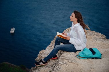 Çekici bir kadın, deniz manzaralı bir dağda oturmuş kitap okuyor. Açık havada kendini eğitmek. günlüğe notlar yazmak,