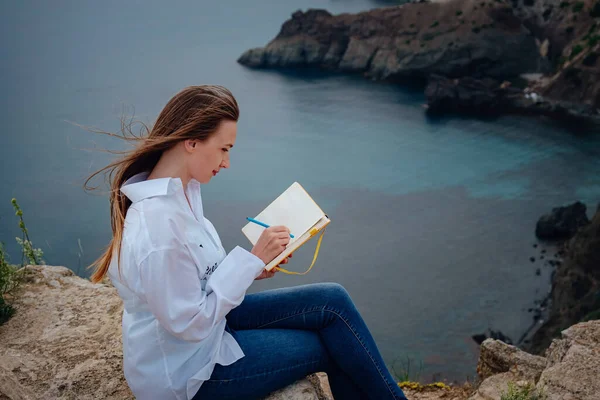 Attractive woman is sitting on the edge of the mountain with a sea view, reading a book. Self-education in the open air. writing notes in diary,